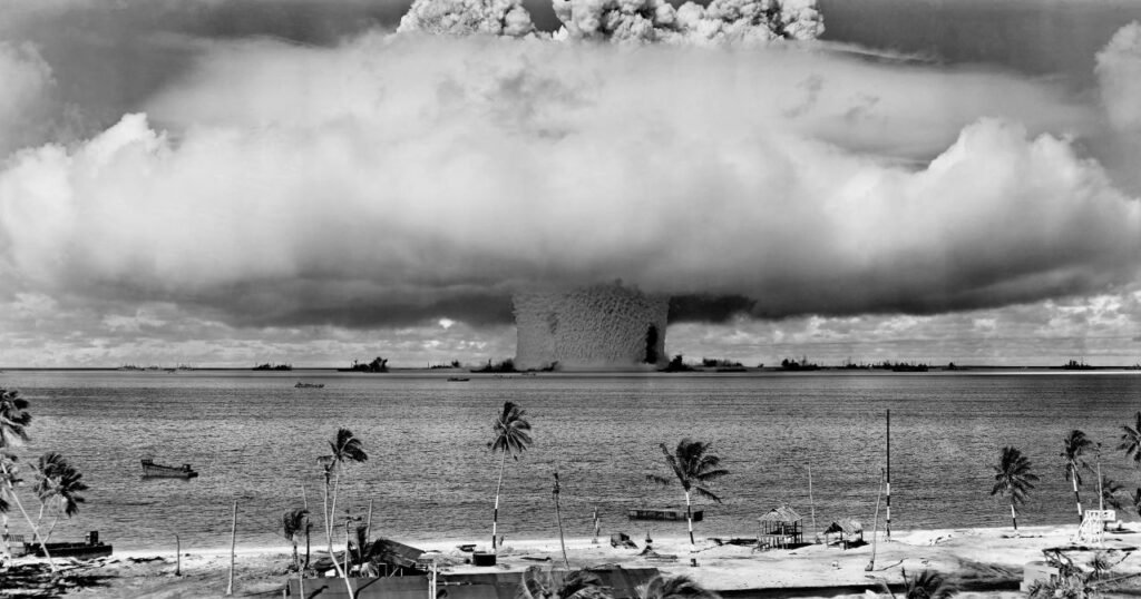 a black and white image of a nuclear explosion seen from a tropical beach with coconut trees marshall islands